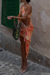 Side view of a model in an orange and yellow patterned mini dress with draped detailing and a matching bra top, holding a textured green bag.