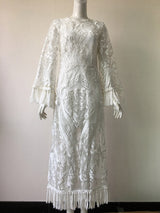 white lace dress on a mannequin, featuring long bell sleeves with fringe, a round neckline, and a midi length with a slit and fringe hem
