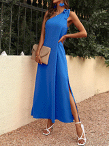 Woman wearing a blue one-shoulder maxi dress with a bow detail on the shoulder and a high slit, holding a beige clutch.