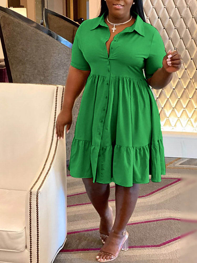 Woman in a green button-down dress with a tiered hem, standing indoors with a patterned wall in the background.