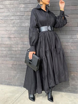 Woman wearing a black tiered maxi dress with balloon sleeves and a wide black belt, holding a black clutch bag.