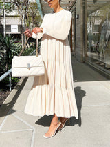 Woman holding a white quilted handbag while wearing a long, cream-colored tiered maxi dress with long sleeves.
