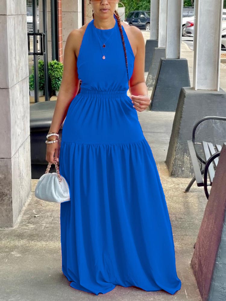 Woman wearing a blue halter neck maxi dress with an elasticated waist and tiered skirt, holding a white clutch bag.