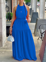 Woman wearing a blue halter neck maxi dress with an elasticated waist and tiered skirt, holding a white clutch bag.