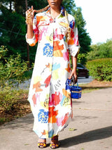 Woman wearing a long, white, collared shirt dress with a colorful abstract print, featuring rolled-up sleeves, a button-down front, and a high-low hemline. She is accessorized with high heels and a small blue handbag.