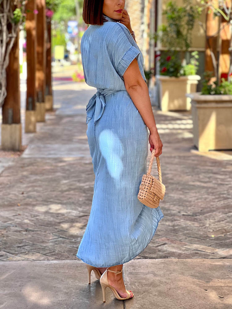 Side view of a woman wearing a light blue denim-like midi dress with short sleeves and a collared neckline. The dress features a self-tie waist and ruched detailing. She is holding a woven beige handbag and wearing nude strappy heels.