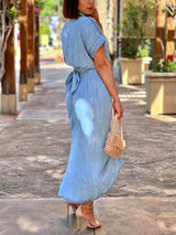 Side view of a woman wearing a light blue denim-like midi dress with short sleeves and a collared neckline. The dress features a self-tie waist and ruched detailing. She is holding a woven beige handbag and wearing nude strappy heels.
