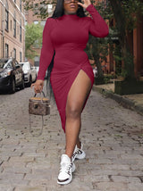 Woman wearing a long-sleeved, high-neck, draped red midi dress with a high slit, walking on a cobblestone street with a handbag.