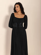 Woman wearing a black midi dress with a square neckline, long puff sleeves, and a corset-style waist detail, viewed from the side with her hands behind her back.