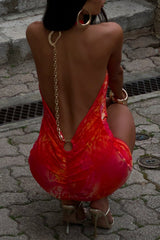 Back view of a woman wearing a red and orange printed mini dress with an open back and chain embellishment.