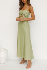 full length shot of a woman in a light green strapless satin dress with a twisted bust and cut-out waist, standing against a white wall