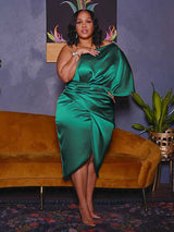 Woman wearing a green one-shoulder satin dress with draping and a tulip hem, standing next to a mustard-colored sofa.