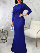 Woman wearing a long royal blue asymmetrical dress with long sleeves and a mermaid hemline, holding a clutch.