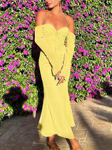 Full body shot of a woman in a yellow ruched halter neck dress with sheer off-the-shoulder sleeves and a flared hem, standing outdoors with a floral background.