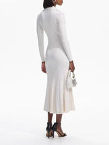 Back view of a woman wearing a cream-colored long-sleeved knit dress with a collared neckline, button-up front, dual chest pockets, and a flared hem, holding a white handbag and wearing black heels.