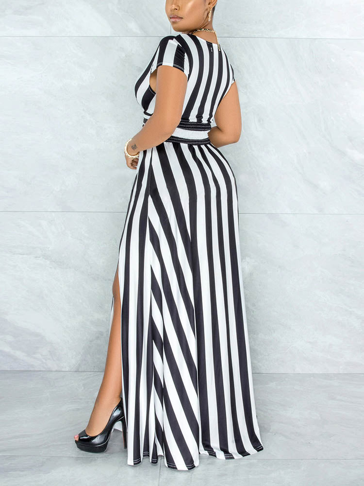 Rear view of a woman wearing a black and white striped maxi dress with a V-neck and high slit, standing against a marble wall.