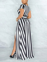 Rear view of a woman wearing a black and white striped maxi dress with a V-neck and high slit, standing against a marble wall.