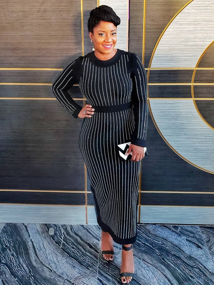 Woman in a black long-sleeved midi dress with vertical rhinestone embellishments, posing with one hand on her hip. She is wearing black open-toe heels and holding a black and white clutch.