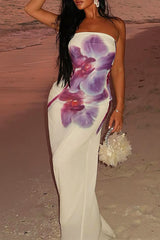 Woman with long dark hair wearing a white strapless maxi dress with a purple floral print, holding a fluffy white handbag on a beach.