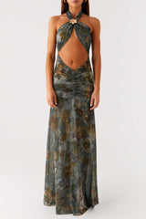 Woman wearing a teal and brown floral print halter neck maxi dress with a cut-out at the midriff, standing against a white wall.