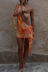 Model wearing an orange and yellow patterned mini dress with a draped overlay, standing against a textured wall.
