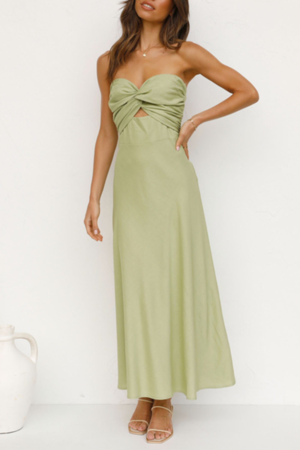 woman with hand on hip in a light green strapless satin dress with a twisted bust and cut-out waist, standing against a white wall