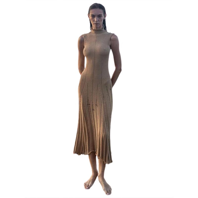 Woman wearing a beige sleeveless ribbed knit midi dress with sheer panel detailing standing on rocks by the ocean against a white background