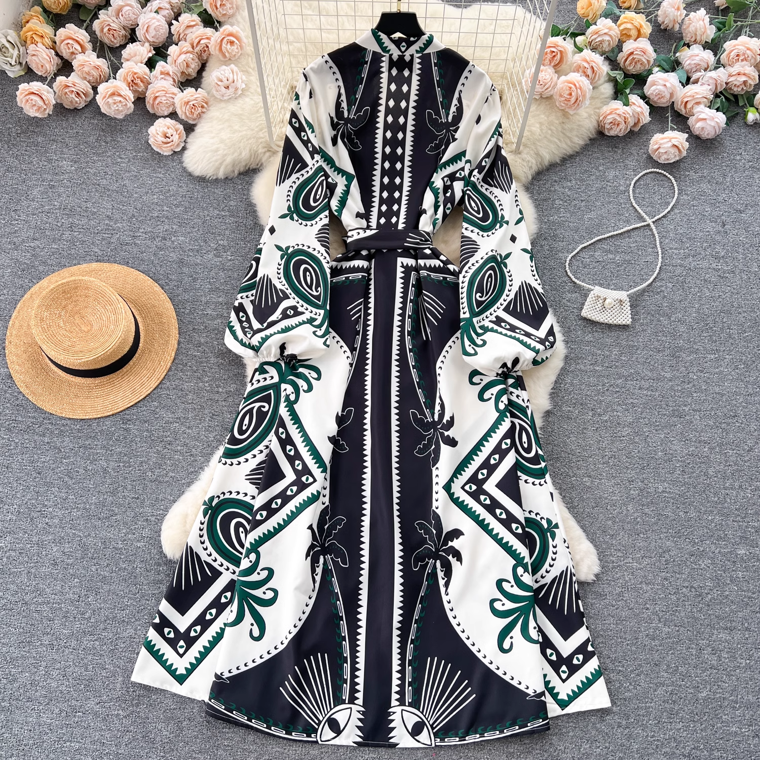 A bohemian print maxi dress with balloon sleeves and a high neckline, laid flat on a white fluffy surface with decorative flowers and a straw hat.