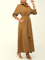 Woman wearing a long brown collared dress with a belted waist, showing pockets, and brown high heels.