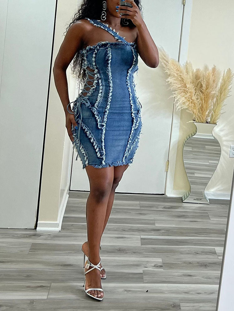 Woman in a blue denim sheath dress with asymmetrical neckline, distressed detailing, and white strappy heels, holding a phone.