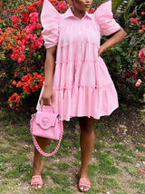Woman in a pink tiered puff sleeve collared dress and pink handbag standing in front of a flowering bush.