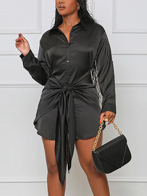 Woman wearing a black satin shirt dress with a tie waist and long sleeves.