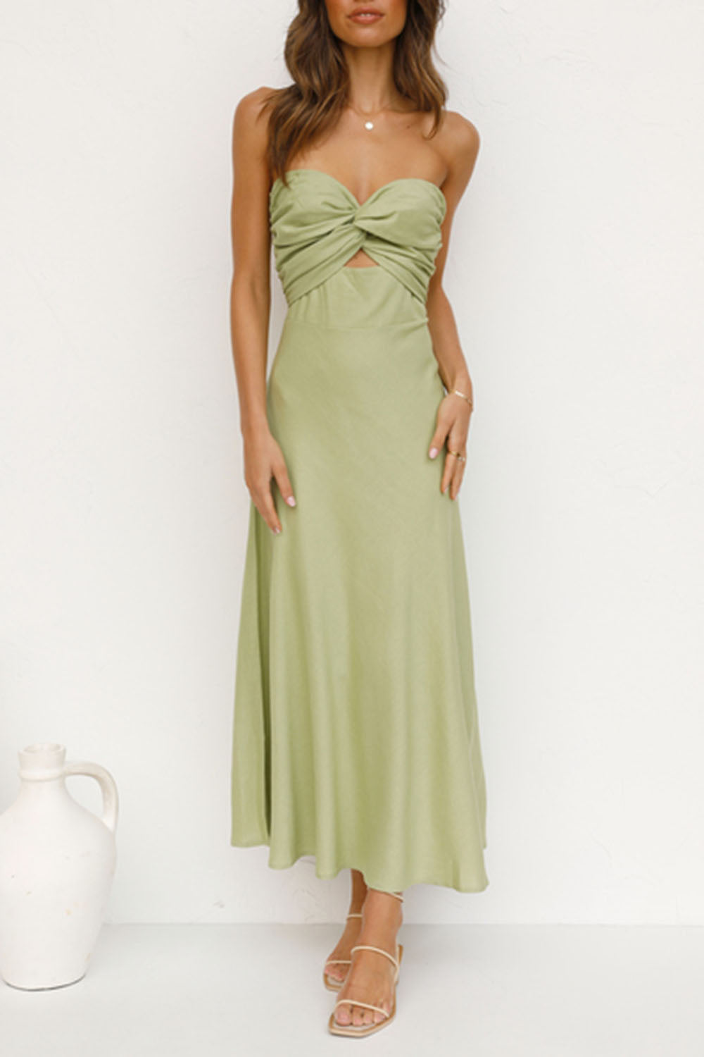 woman posing in a light green strapless satin dress with a twisted bust and cut-out waist, standing against a white wall