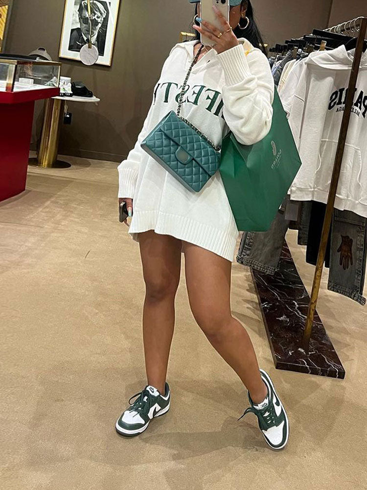 Woman wearing a white oversized knit sweater dress with green 'LIFESTYLE' text graphic, a green quilted handbag, and a green shopping bag, standing in a store
