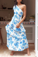 Woman in a white and blue floral print one-shoulder tiered maxi dress, lifting the hem of the skirt.