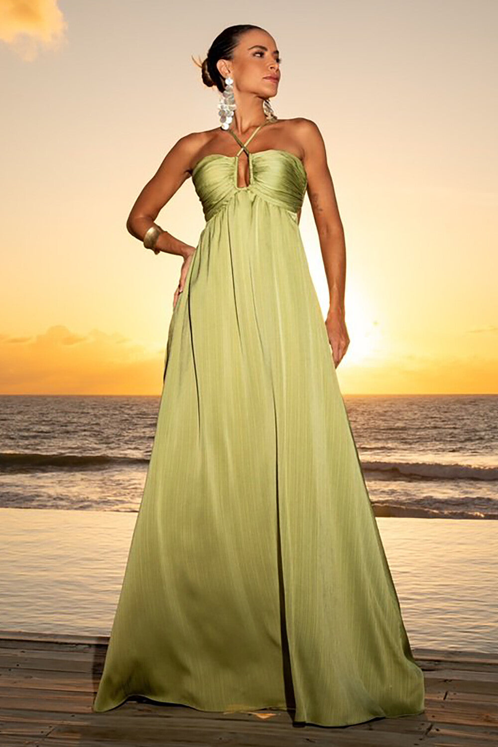 Woman wearing a flowing lime green halter maxi dress with an open back, standing on a wooden deck by the ocean at sunset.