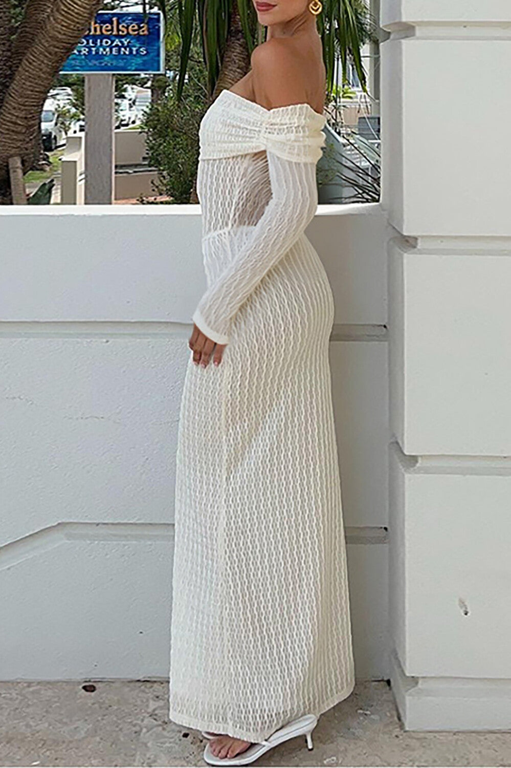 Side view of a woman wearing a white off-the-shoulder maxi dress with long sleeves and a textured knit overlay, standing outdoors.