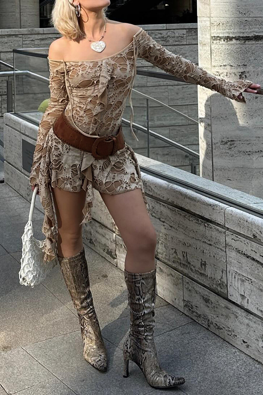 A woman poses in a beige distressed off-the-shoulder dress, brown belt, and snakeskin boots.