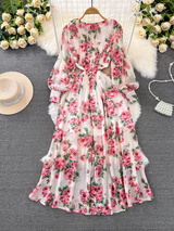 A white chiffon maxi dress with a pink rose print, shown hanging on a garment rack with a white faux fur rug and decorative flowers in the background.