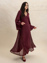 Woman wearing a burgundy maxi dress with flared sleeves and lace inserts, posing with her hand outstretched.