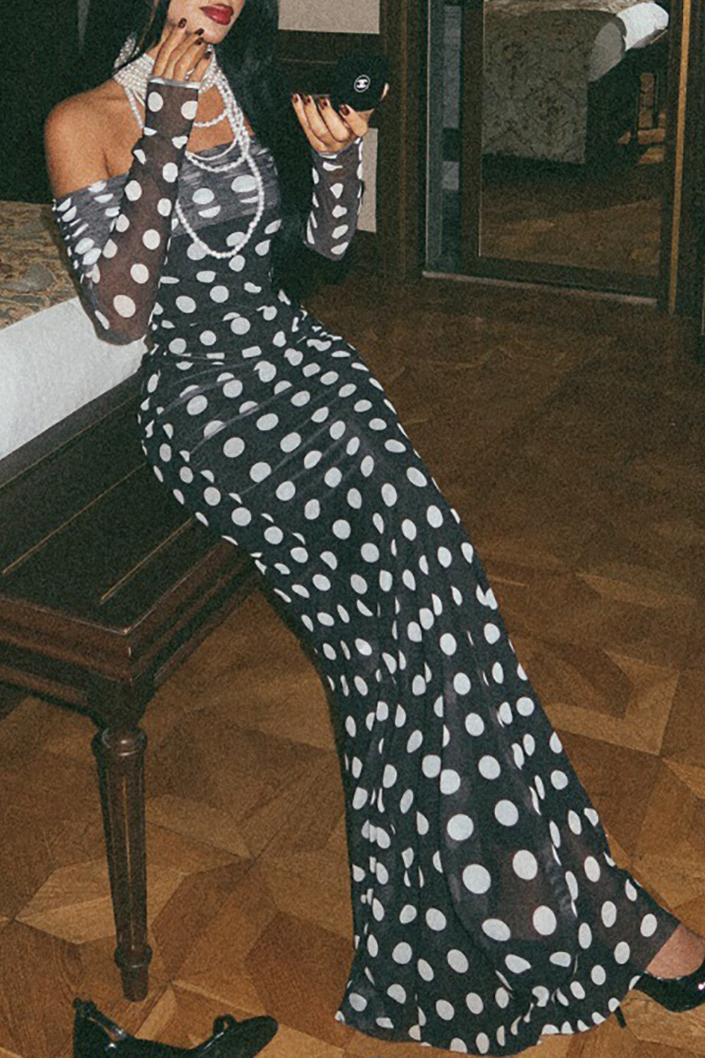 Close-up of a woman wearing a black and white polka dot off-the-shoulder maxi dress with long sleeves, sitting on a wooden bench and looking at a compact.