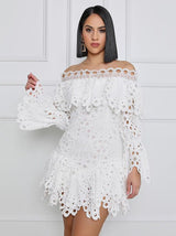 Woman wearing a white off-the-shoulder eyelet lace mini dress with bell sleeves and ruffled tiers, standing against a white wall.