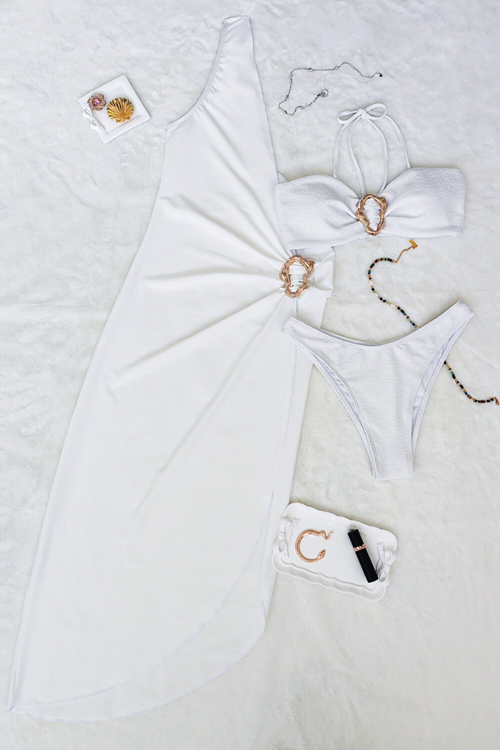 Flat lay of a white one-shoulder maxi dress with a matching bikini set and accessories.