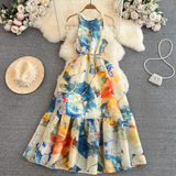 Overhead view of a colorful abstract floral print maxi dress with a tiered skirt and tie waist, laid out with a straw hat and a pearl beaded handbag.
