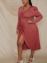 Plus-size woman in a rust-colored long-sleeved V-neck ruched wrap dress with puff sleeves, with one hand on her hip against a beige backdrop.