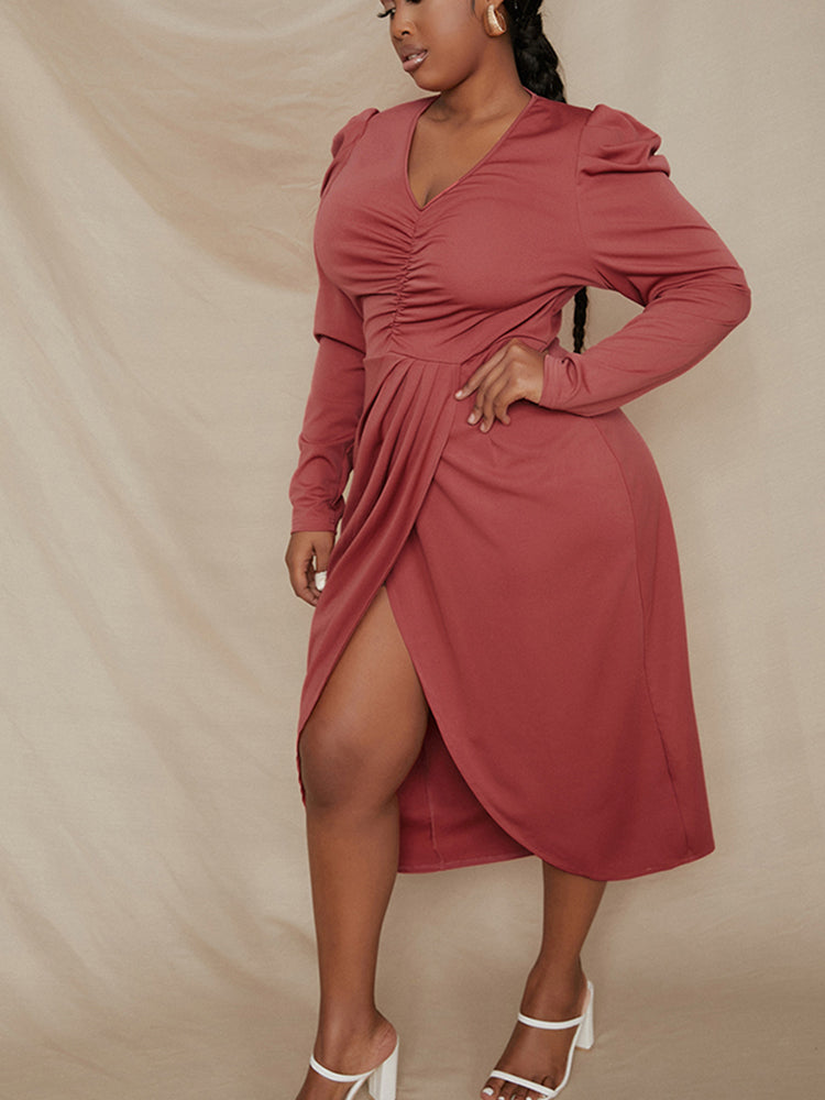Plus-size woman in a rust-colored long-sleeved V-neck ruched wrap dress with puff sleeves, with one hand on her hip against a beige backdrop.