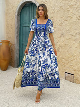 Woman wearing a blue and white floral print maxi dress with puff sleeves and a square neckline, holding a straw bag.