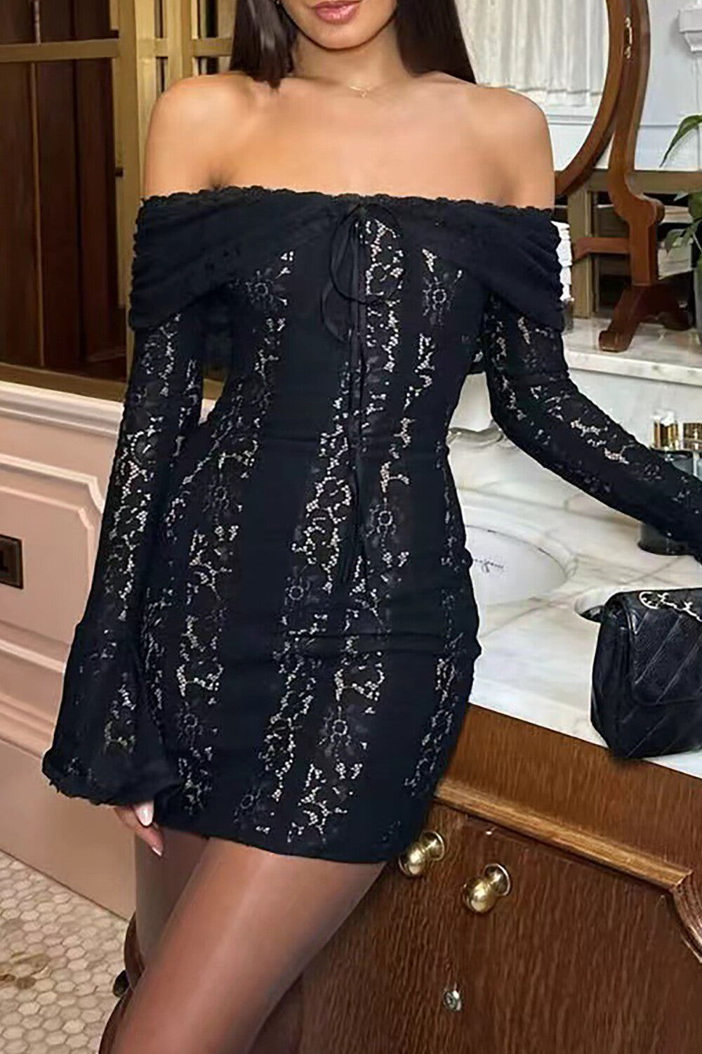 Close-up of a woman wearing a black off-the-shoulder lace mini dress with long bell sleeves and a tie-front detail, styled with sheer tights.