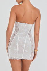 Back view of a woman wearing a white strapless lace mini dress with boning details and scalloped hem, showing the zipper closure.