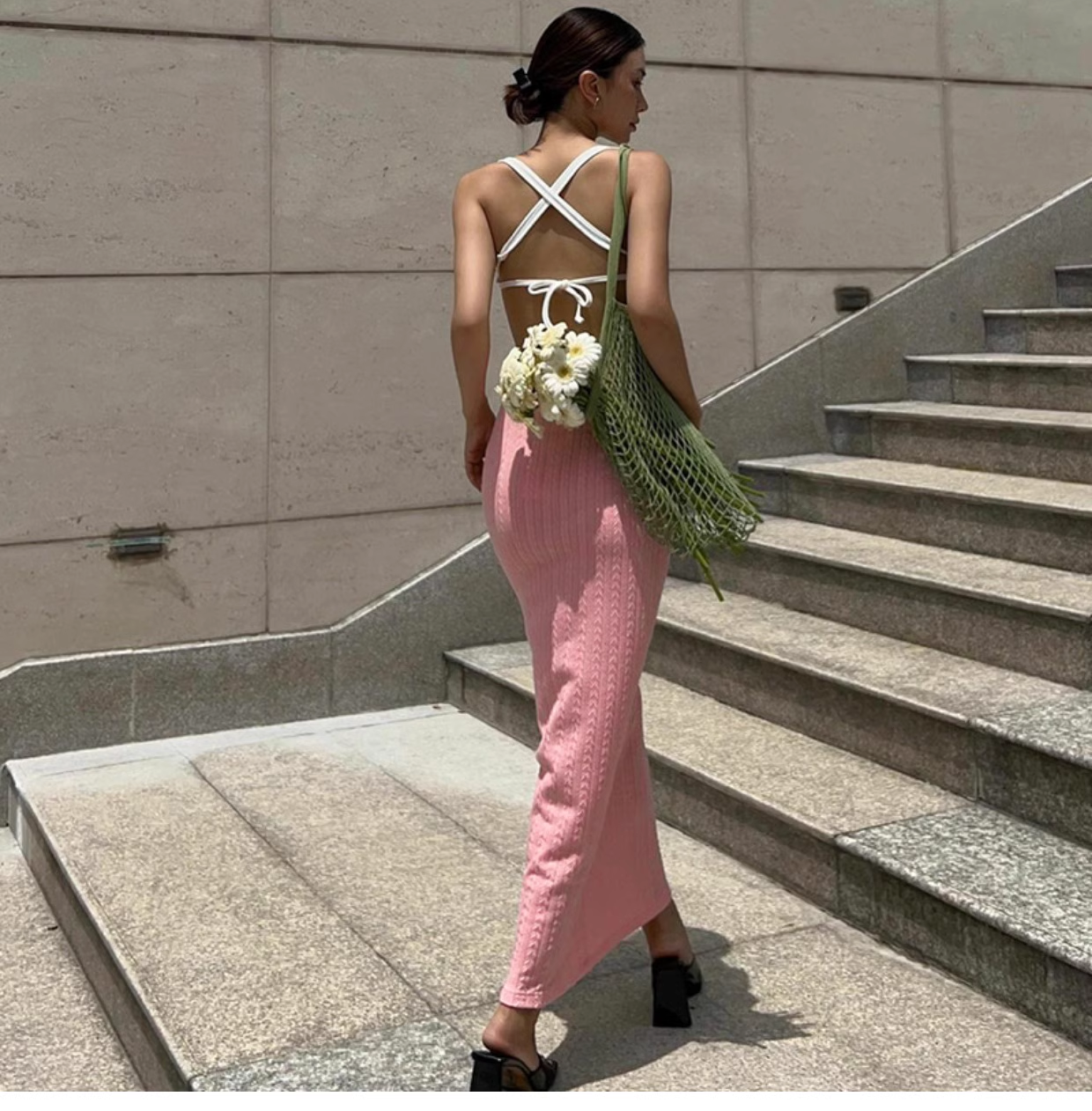 Rear view of a woman wearing a pink knit midi dress with cut-out details and a green mesh bag with flowers, walking up stairs.